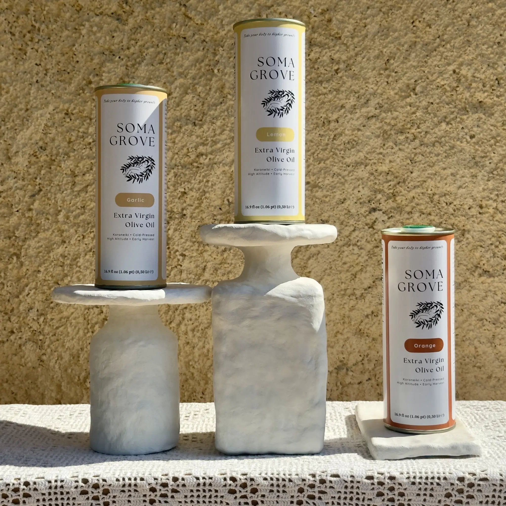 The Soma Grove Flavor Trinity set, featuring Garlic, Lemon, and Orange flavored extra virgin olive oil cans displayed on artistic white pedestals.