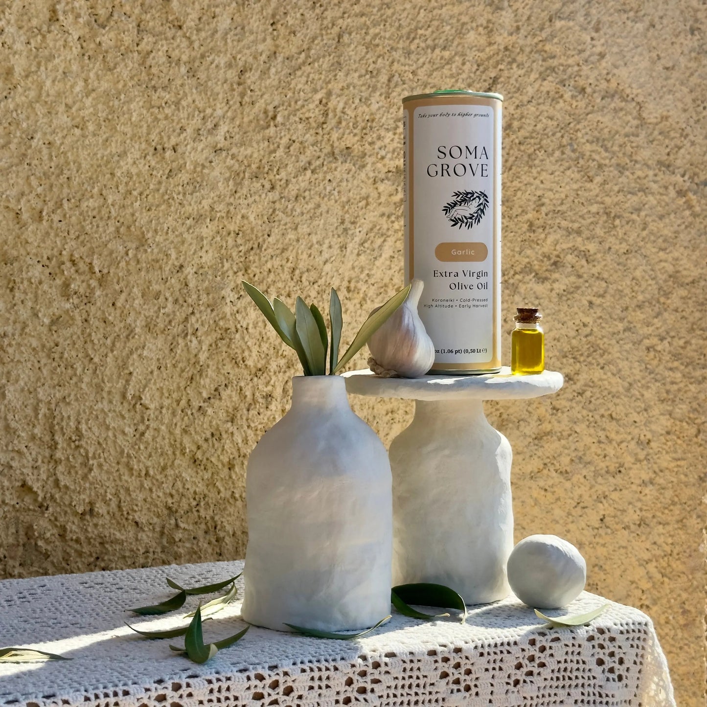 Soma Grove Garlic olive oil can with garlic cloves and olive leaves scattered across traditional Greek textiles.