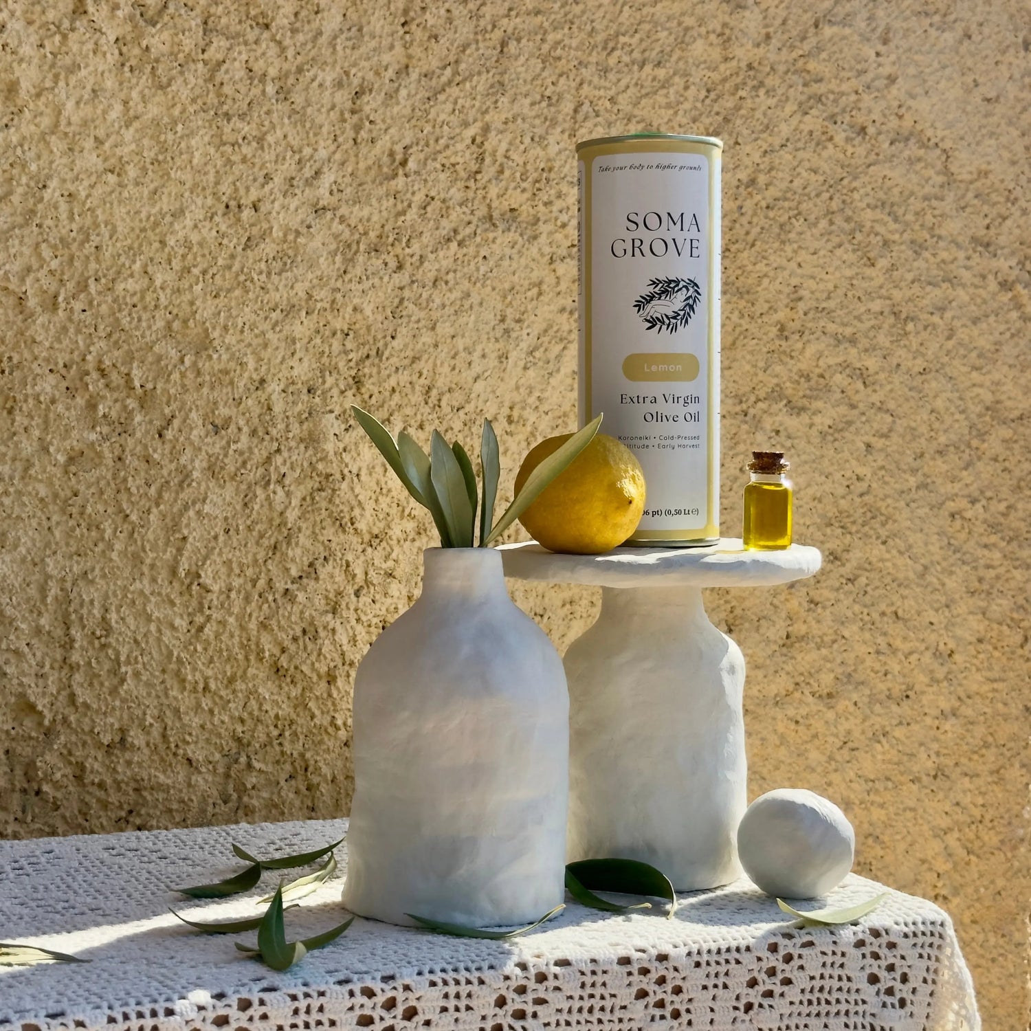Soma Grove Lemon olive oil can with a lemon and olive leaves scattered across traditional Greek textiles.